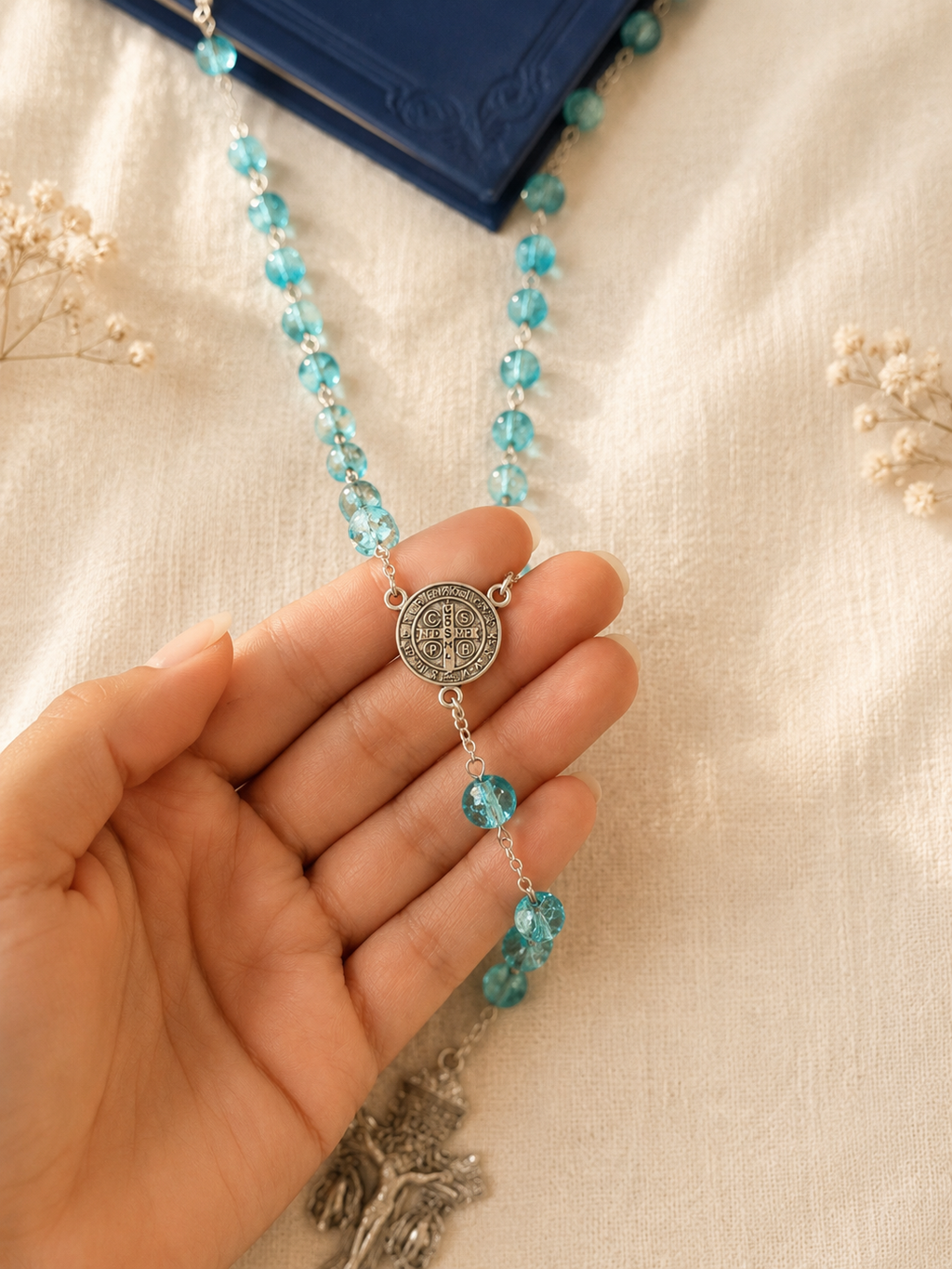 Turquoise cristal beaded rosary with a silver pendant, Saint Benedict's medal and the 3-way Pardon Indulgence Crucifix held by hand on a beige fabric background