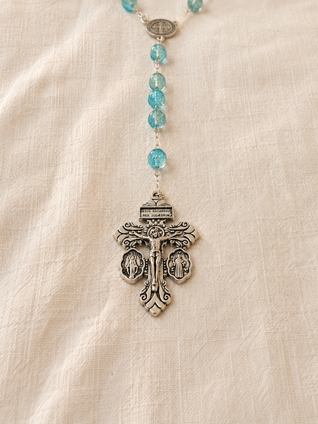 Turquoise cristal beaded rosary with a silver pendant, Saint Benedict's medal and the 3-way Pardon Indulgence Crucifix on a beige fabric background